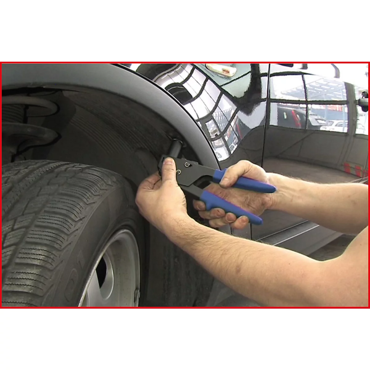 Person repariert Kotflügel. Hände halten Zange mit blauen Griffen. Reifen im Vordergrund, schwarzes Auto.